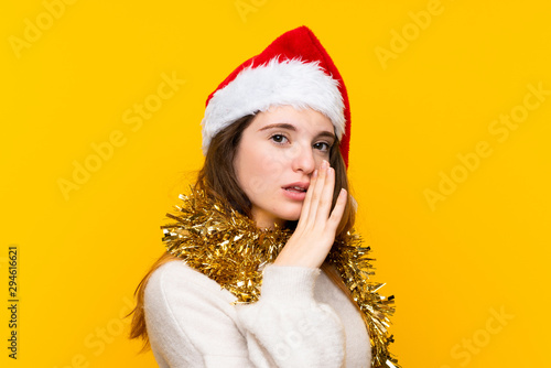 Girl with christmas hat over isolated yellow background whispering something