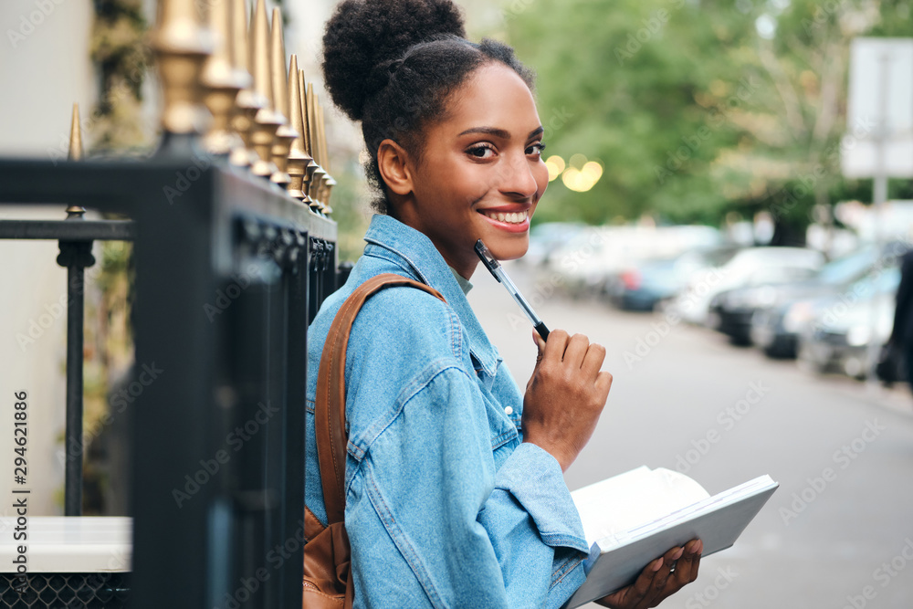 Cheerful casual African American student girl in denim jacket with notepad happily looking in camera on street Cheerful casual African American student girl in denim jacket with notepad happily looking in camera on street