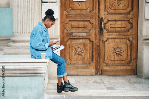 Side view of casual African American student girl in denim jacket with cellphone and book thoughtfully studying on street