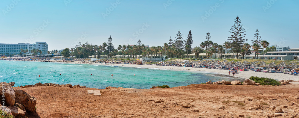 Fototapeta premium Panoramic shot of a beach with stones and transparent water of the azure mediterranean sea, surrounded by a picturesque nature of Cyprus. Ayia Napa.