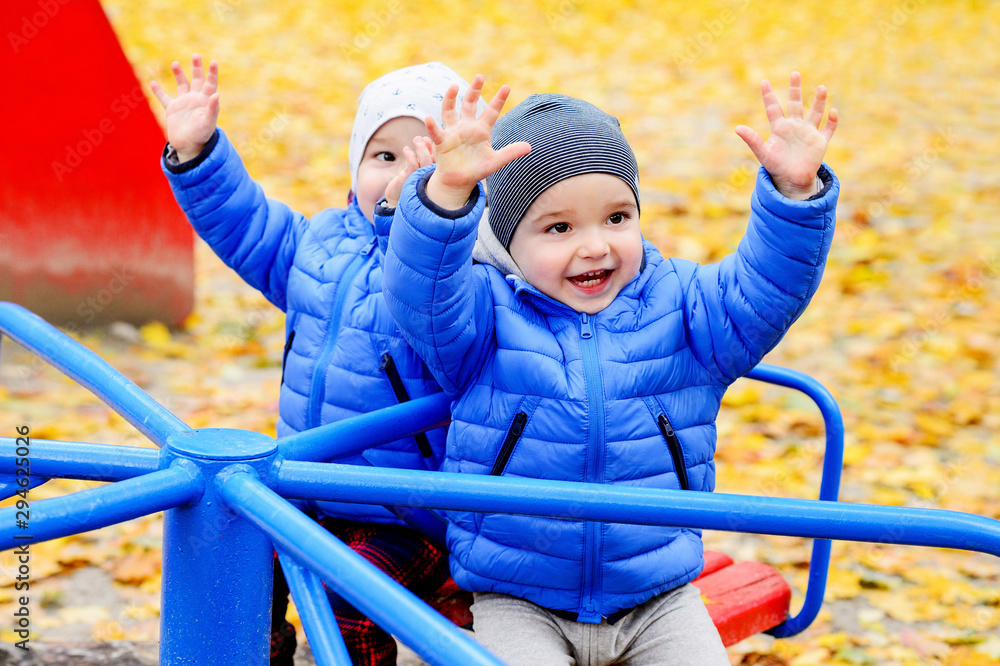 Brightly dressed twin brothers play on a carousel in an autumn park amid fallen yellow leaves. They are two years old. Brightly dressed twin brothers play on a carousel in an autumn park amid fallen yellow leaves. They are two years old.