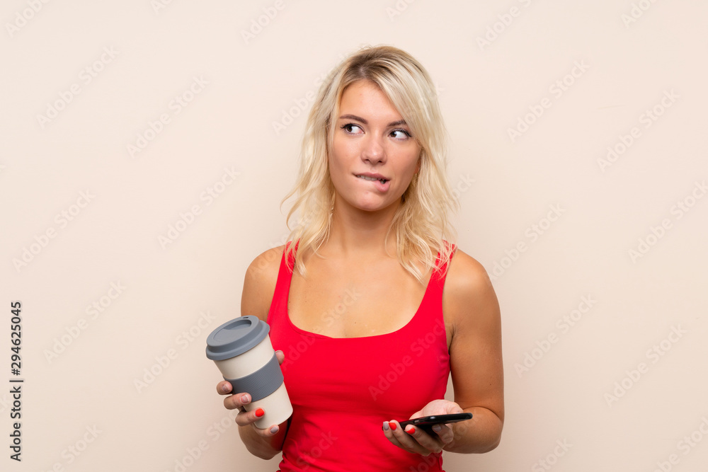 Young blonde woman over isolated background holding coffee to take away and a mobile while thinking something Young blonde woman over isolated background holding coffee to take away and a mobile while thinking something