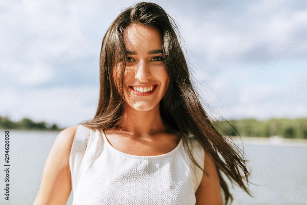 Happy beautiful young woman smiling broadly with healthy toothy smile and with a windy blowing long hair during walking in the park, posing on nature background.