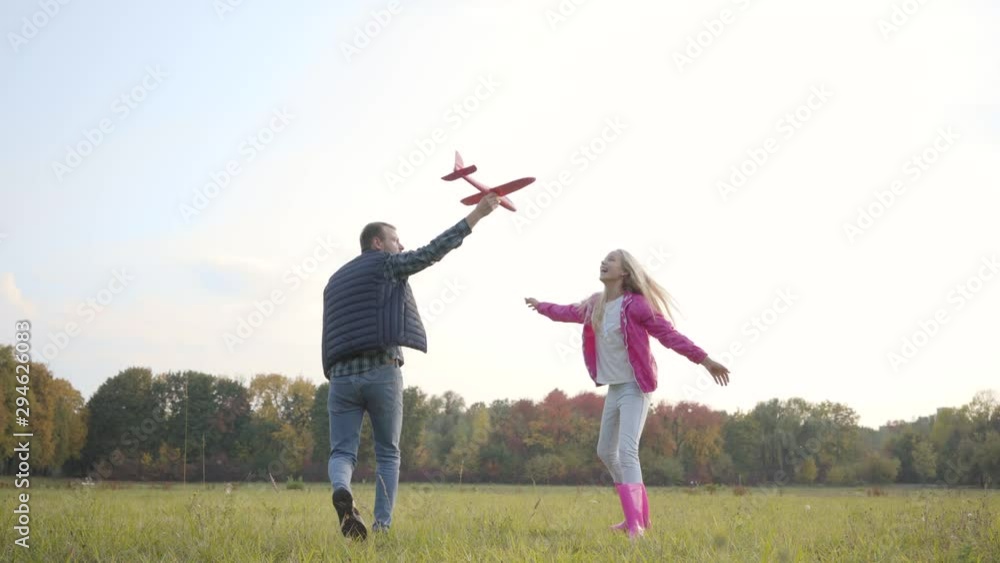 Back view of caucasian man and young blonde girl strolling and spinning on the autumn meadow. Father holding pink toy airplane imitating its fly and his daughter running next to him and laughing.