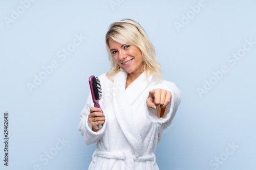 Young woman in a bathrobe with hair comb points finger at you with a confident expression