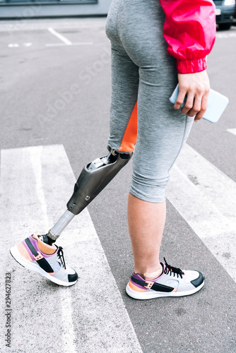 partial view of disabled sportswoman with prosthesis holding smartphone on street