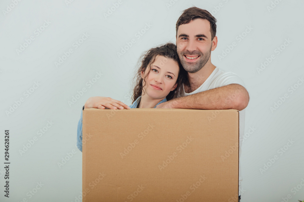 Smiling wife and husband standing behind the big carton box, together, hugging, looking at the camera. Smiling wife and husband standing behind the big carton box, together, hugging, looking at the camera.