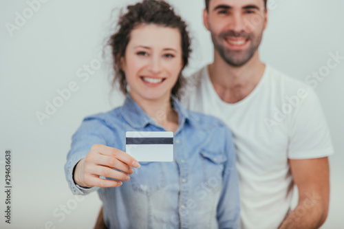 Blurred husband is hugging his wife. Pretty wife is holding a credit card in front of the camera. They are smiling. Focus on the card.