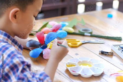 Asian boy painting a colourful plastic flower made from water bottle on wooden table outdoor.Recycled colorful plastic flowers.Recycle decoration.