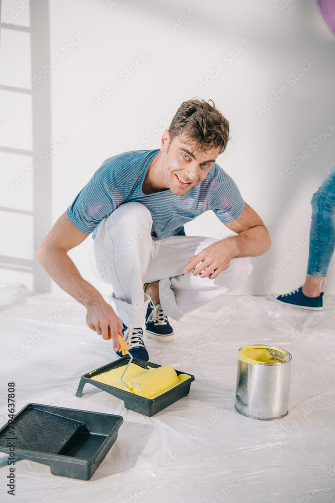 handsome young man putting paint roller into roller tray with yellow paint and smiling at camera handsome young man putting paint roller into roller tray with yellow paint and smiling at camera