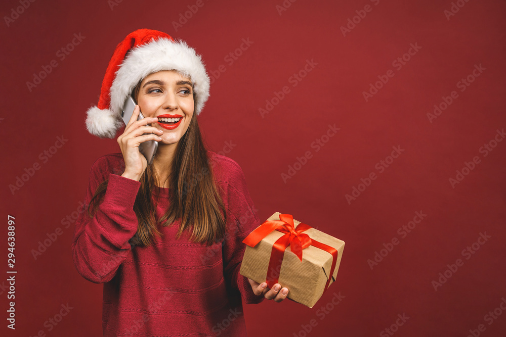 Smiling girl with Christmas gifts calling on phone. Santa's helper. Girl with Santa's hat isolated against red background. Using phone. Smiling girl with Christmas gifts calling on phone. Santa's helper. Girl with Santa's hat isolated against red background. Using phone.