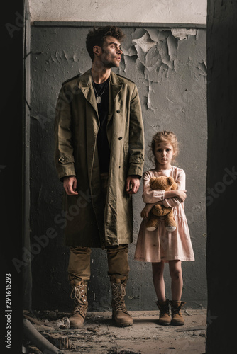 handsome man standing near kid with teddy bear near weathered wall, post apocalyptic concept
