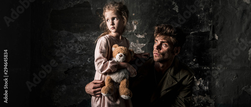 panoramic shot of upset man near kid with teddy bear in dirty room, post apocalyptic concept