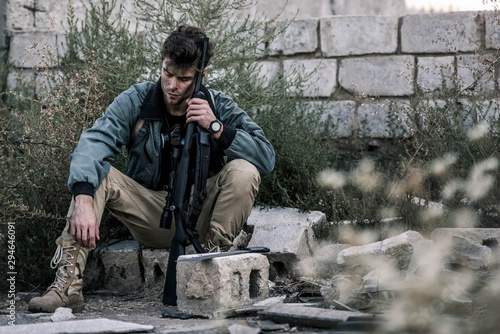 selective focus of handsome man sitting with gun near brick wall outside, post apocalyptic concept © LIGHTFIELD STUDIOS