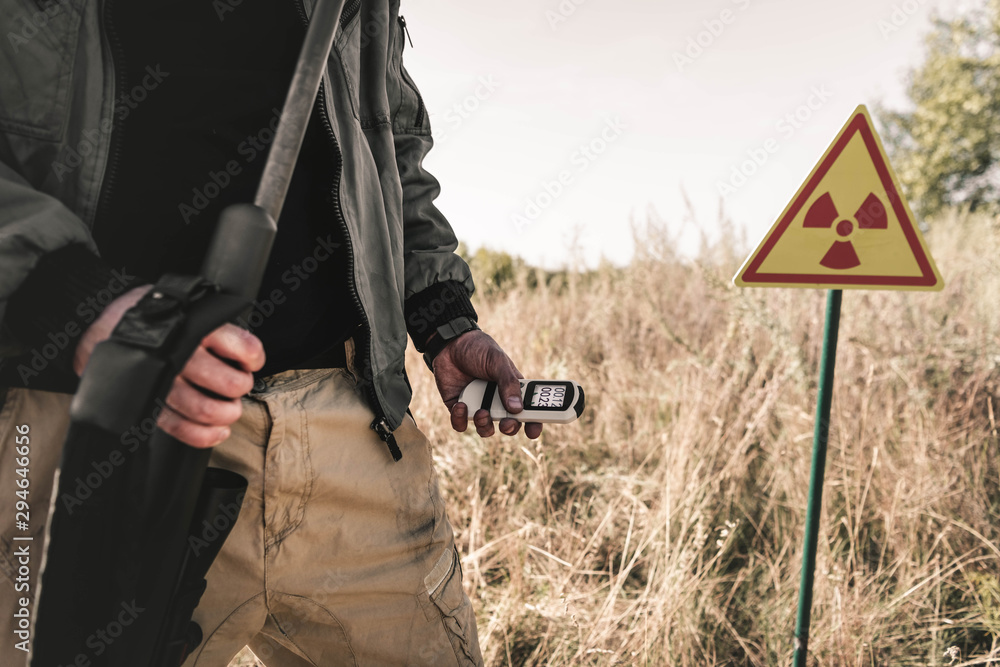 cropped view of man holding radiometer and gun near toxic symbol, post apocalyptic concept