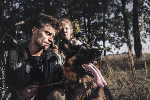 selective focus of handsome man holding leash of german shepherd dog near kid, post apocalyptic concept