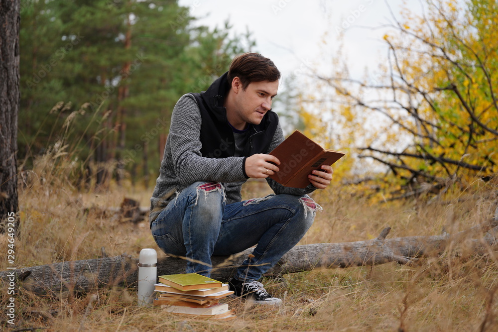 Fototapeta premium A young handsome man in casual clothes is sitting on a log and reading a book in the autumn forest.