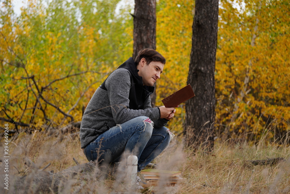 A young handsome man in casual clothes is sitting on a log and reading a book in the autumn forest. A young handsome man in casual clothes is sitting on a log and reading a book in the autumn forest.