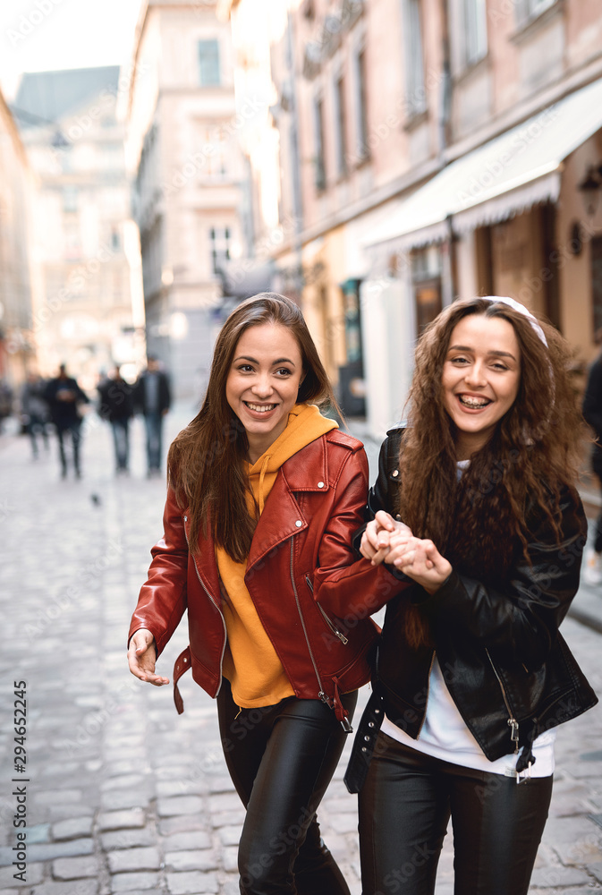 portrait of young female friends in colored leather biker jackets and hoodies holding hands while walking down the street.