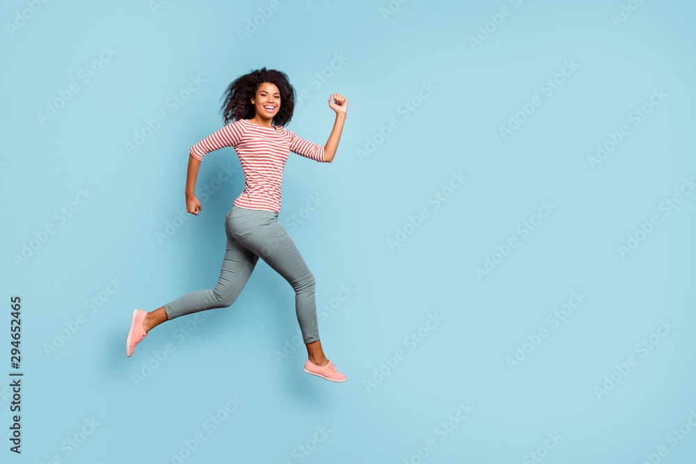 Full length body size side profile photo of cheerful excited cute nice curly wavy youngster running jumping for sales wearing trousers pants striped t-shirt sneakers isolated over pastel blue color