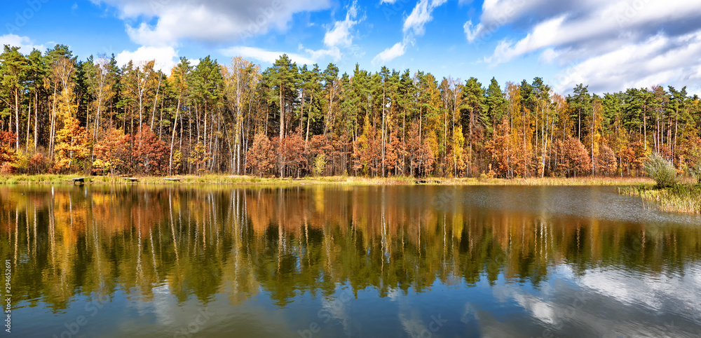 Fototapeta premium golden autumn landscape with forest trees reflecting in lake water against clouds blue sky panorama wide view of yellow red orange green natural color of fall season nature idyllic panoramic wallpaper