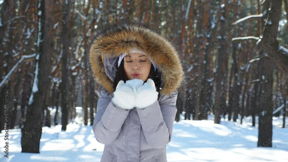Young cheerful girl in white gloves blowing off snow from her palms. Portrait of beautiful woman standing in winter forest and playing with snow. Snowy wood at background. Slow motion Close up Young cheerful girl in white gloves blowing off snow from her palms. Portrait of beautiful woman standing in winter forest and playing with snow. Snowy wood at background. Slow motion Close up