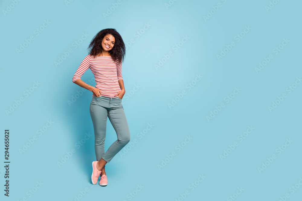 Full length photo of pretty dark skin lady adorable smile holding hands in pockets glad sunny day weather wear casual clothes isolated pastel blue color background