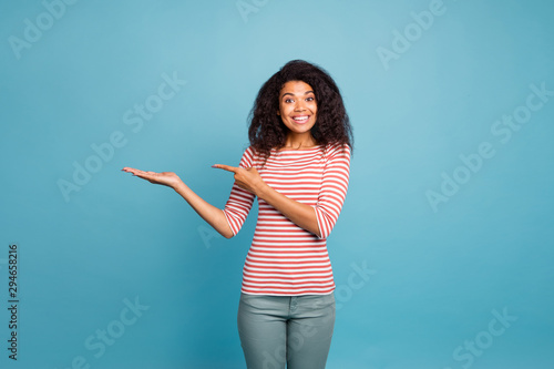 Photo of cheerful excited ecstatic crazy cheerful nice cute sweet pretty black girlfriend pointing at empty space on her palm smiling toothily wearing grey trousers pants isolated over blue vivid