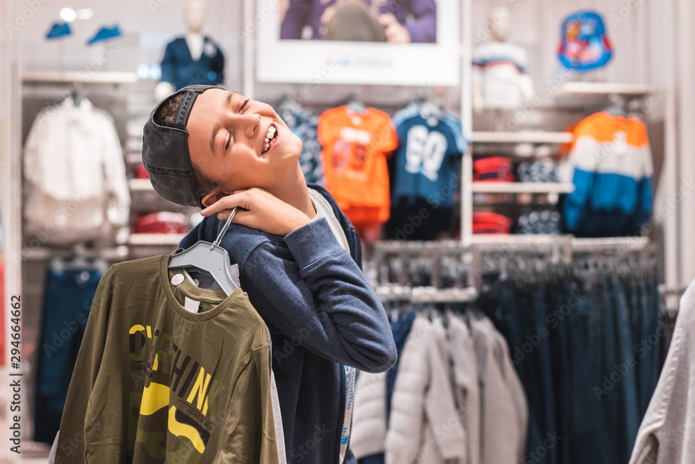 Young boy choosing clothes in the shop by himself, putting on new looks, checking for style. Shopping, black friday, discount and sales concept.