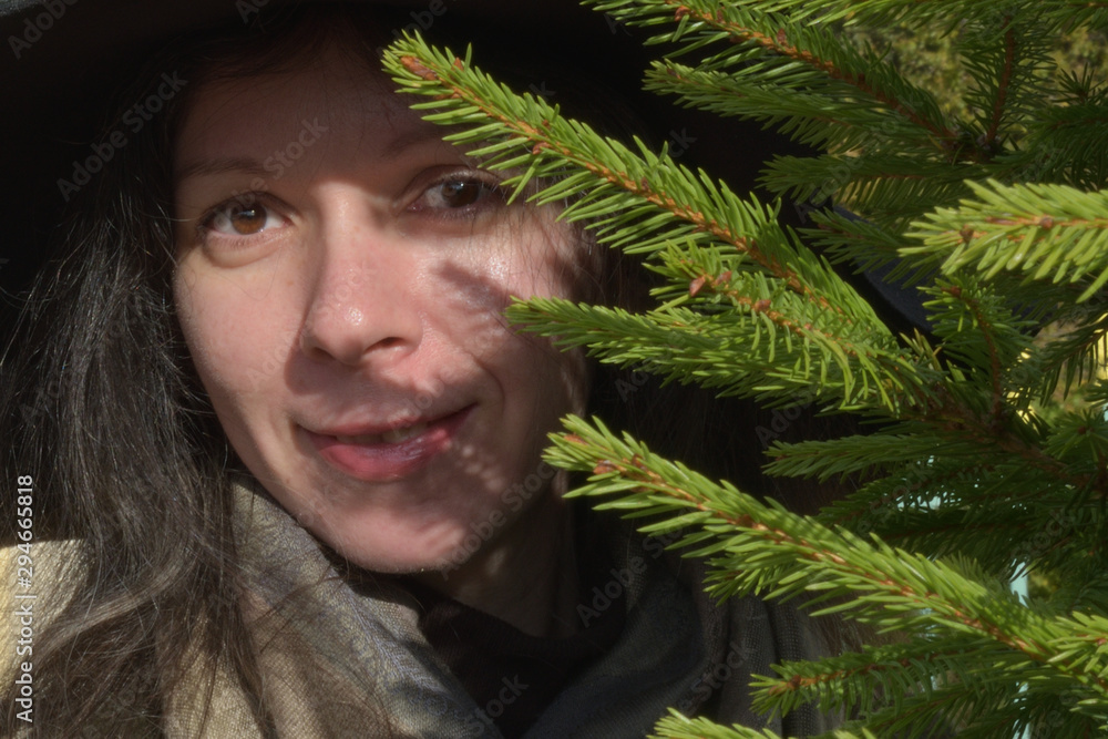 beautiful young lady girl in black hat and green and brown overcoat looking from behind a fir tree smiling beautiful young lady girl in black hat and green and brown overcoat looking from behind a fir tree smiling