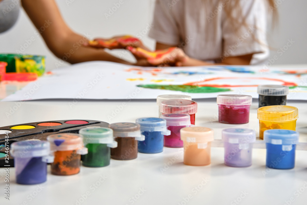 Little artist in white t-shirt standing at table with whatman and colorful paints, painting on it with her hands. Isolated on white. Close-up.