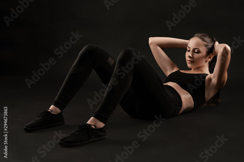 Brunette woman in black leggings, top and sneakers is posing against a black background. Fitness, gym, healthy lifestyle concept. Full length.