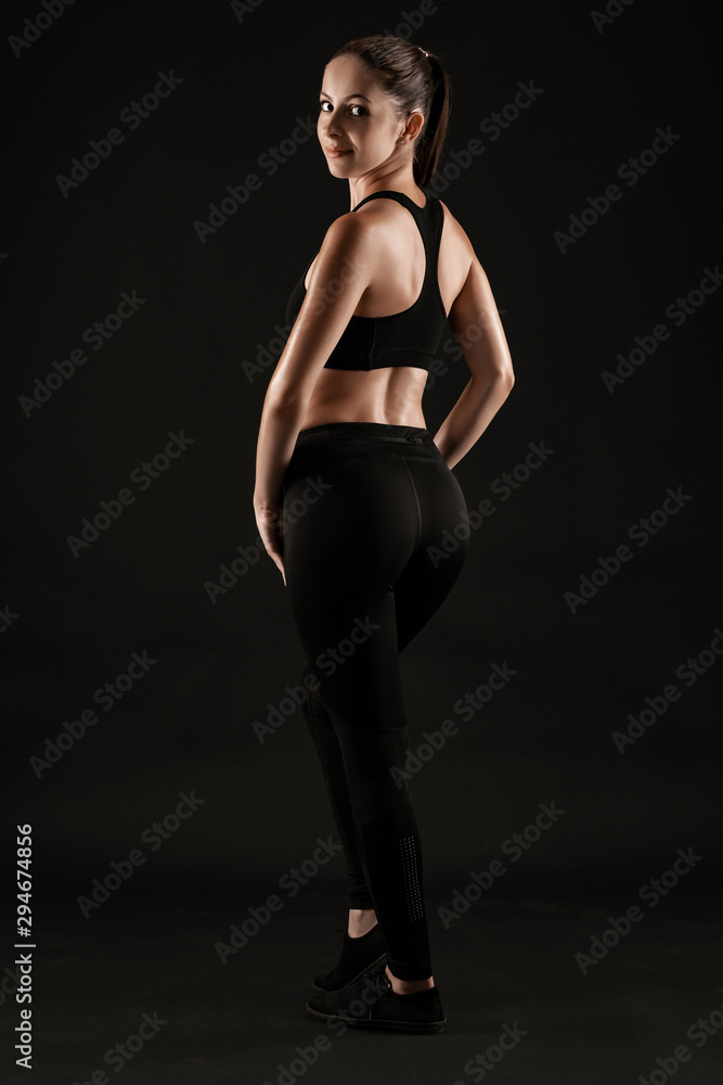 Brunette woman in black leggings, top and sneakers is posing against a black background. Fitness, gym, healthy lifestyle concept. Full length.