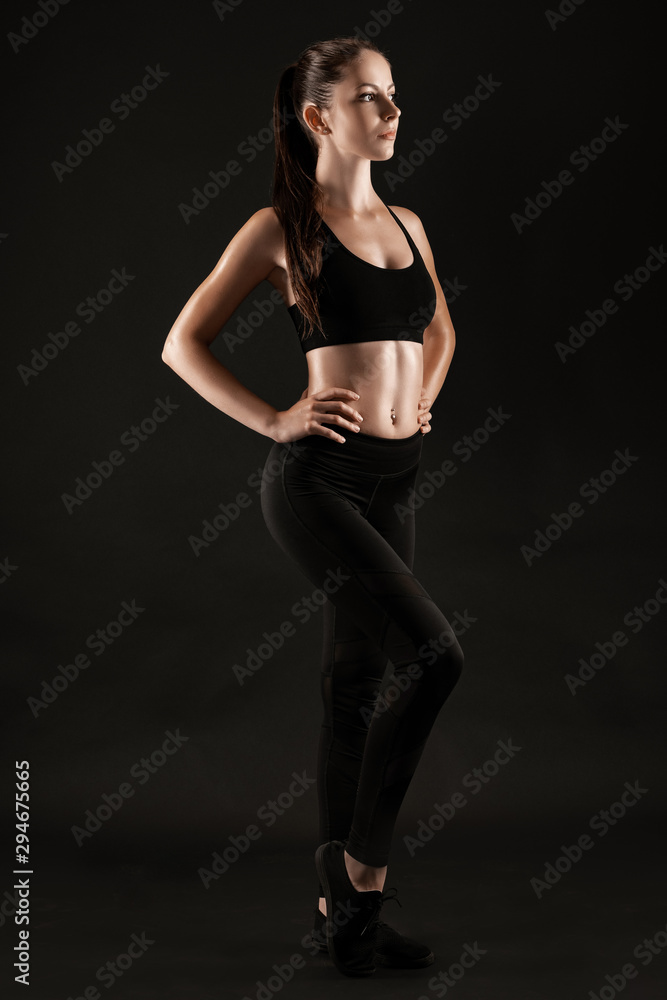 Brunette woman in black leggings, top and sneakers is posing against a black  background. Fitness, gym, healthy lifestyle concept. Full length. Stock  Photo