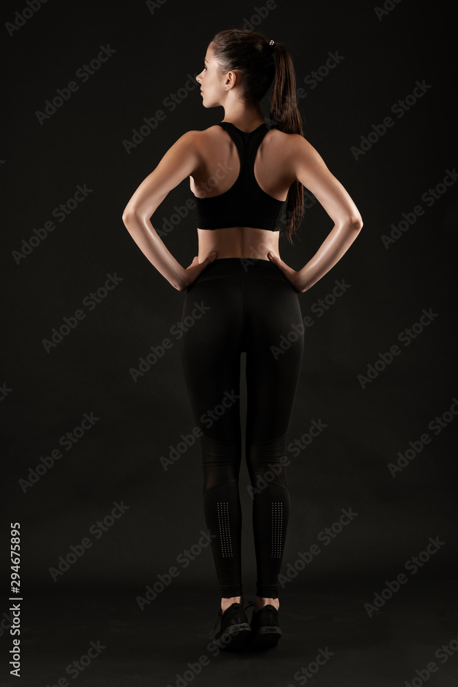Brunette woman in black leggings, top and sneakers is posing against a black background. Fitness, gym, healthy lifestyle concept. Full length.