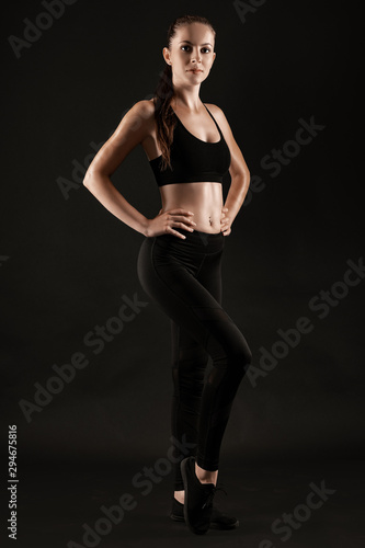 Brunette woman in black leggings, top and sneakers is posing against a black background. Fitness, gym, healthy lifestyle concept. Full length.