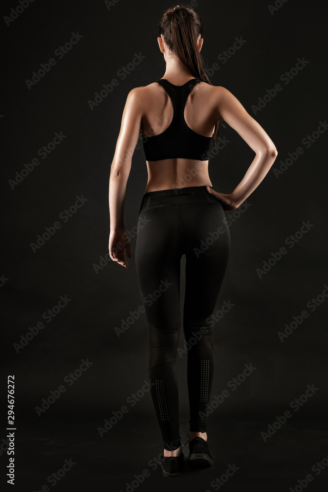 Fototapeta premium Brunette woman in black leggings, top and sneakers is posing against a black background. Fitness, gym, healthy lifestyle concept. Full length.