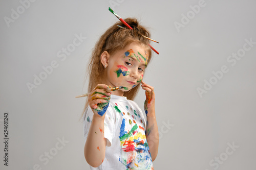 Little girl in white t-shirt, with brushes in her hair is posing standing isolated on white, gesticulating with painted hands and face. Close-up.