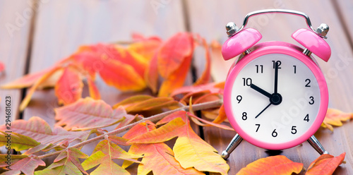Web banner of daylight savings time concept, pink alarm clock with orange autumn leaves on a wooden background