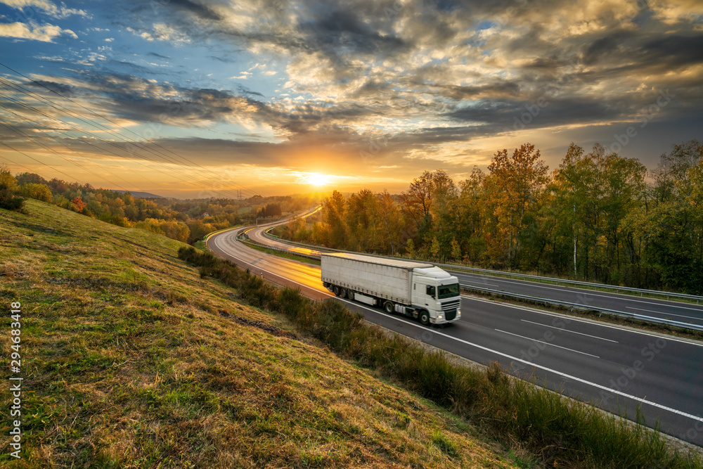 White truck driving on the asphalt highway in autumn landscape at golden sunset with dramatic clouds