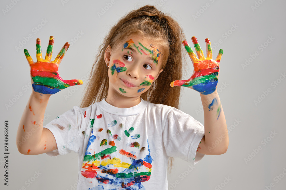 Little girl in white t-shirt is posing standing isolated on white and gesticulating with her painted in different colors palms. Art studio. Close-up. Little girl in white t-shirt is posing standing isolated on white and gesticulating with her painted in different colors palms. Art studio. Close-up.