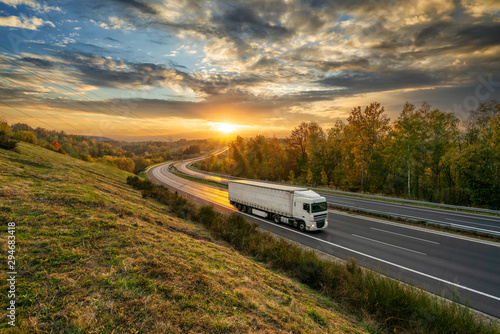 White truck driving on the asphalt highway in autumn landscape at golden sunset with dramatic clouds