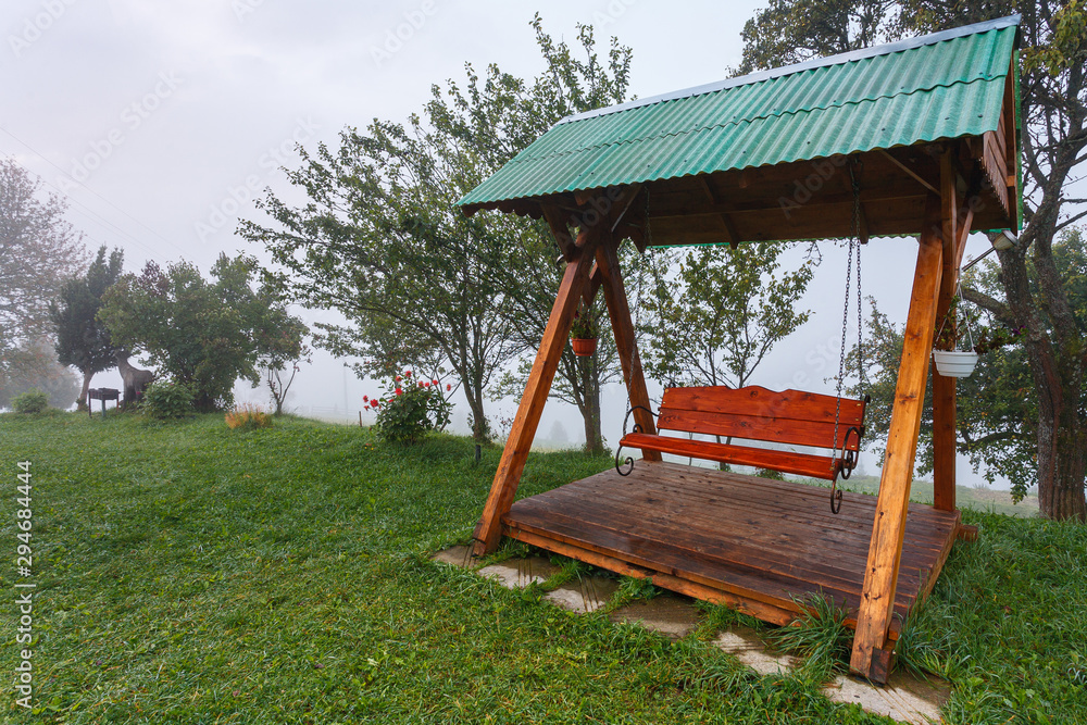 Red swing bench with green roof in forest at foggy autumn morning with trees and vivid green grass Red swing bench with green roof in forest at foggy autumn morning with trees and vivid green grass