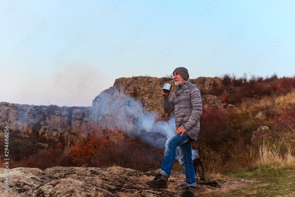 Old gray-haired tourist with a beard drinking hot tea from an iron mug by the fire against the background of the gorge and stones Old gray-haired tourist with a beard drinking hot tea from an iron mug by the fire against the background of the gorge and stones