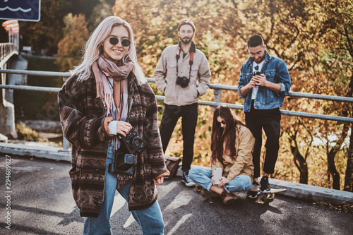 Attractive blond girl with photo camera is enjoying autumn walk with her friends.