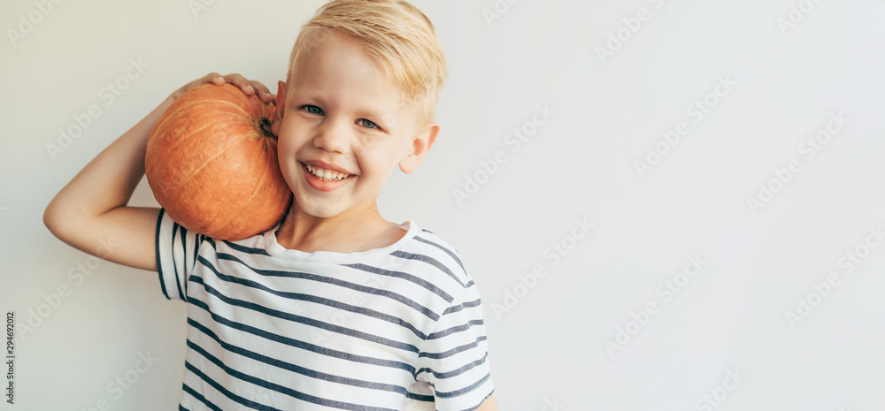 Blond funny Caucasian boy in a striped T-shirt jokes with a ripe pumpkin. Cheerful portrait of a child. Copy space for text. Halloween content.