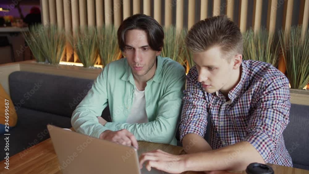 Camera zooming in to young men in casual outfits sitting at restaurant table and fulfilling homework assignment on laptop Camera zooming in to young men in casual outfits sitting at restaurant table and fulfilling homework assignment on laptop