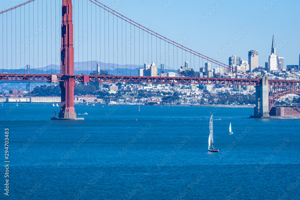Panoramic view of San Francisco and the Golden gate Bridge on a sunny day, with blue water and sailboats in the foreground.