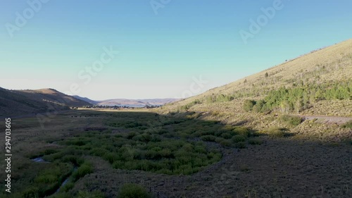 Aerial-Ascending over shadowy mountain meadow with small stream meandering through it with view of small mountain town and lake in the far distance. photo