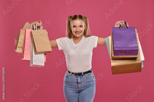 Blonde woman with ponytails, dressed in white t-shirt and jeans posing against a pink background with packages. Close-up. Sincere emotions, shopping.